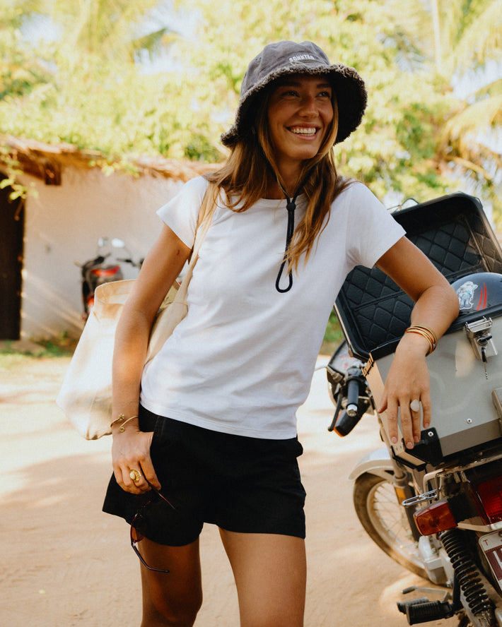 A woman wearing a white crewneck t-shirt, black DUER womens shorts, and a dark gray bucket hat stands next to a motorcycle in an outdoor setting with green foliage in the background.