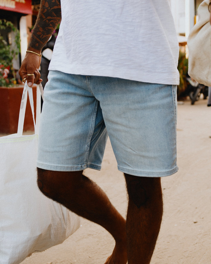 A person wearing light-wash DUER denim shorts and a white t-shirt, carrying a white plastic bag in an outdoor setting.