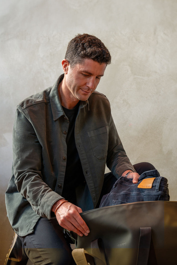Man in a green jacket packing DUER Performance Jeans into a black bag against a plain background