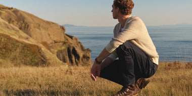 man wearing Duer Jeans sitting on a grassy hill overlooking a body of water with mountains in the background