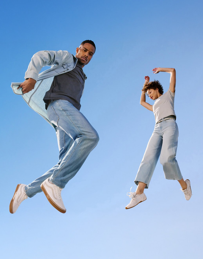 Two people jumping against a clear blue sky wearing DUER clothing