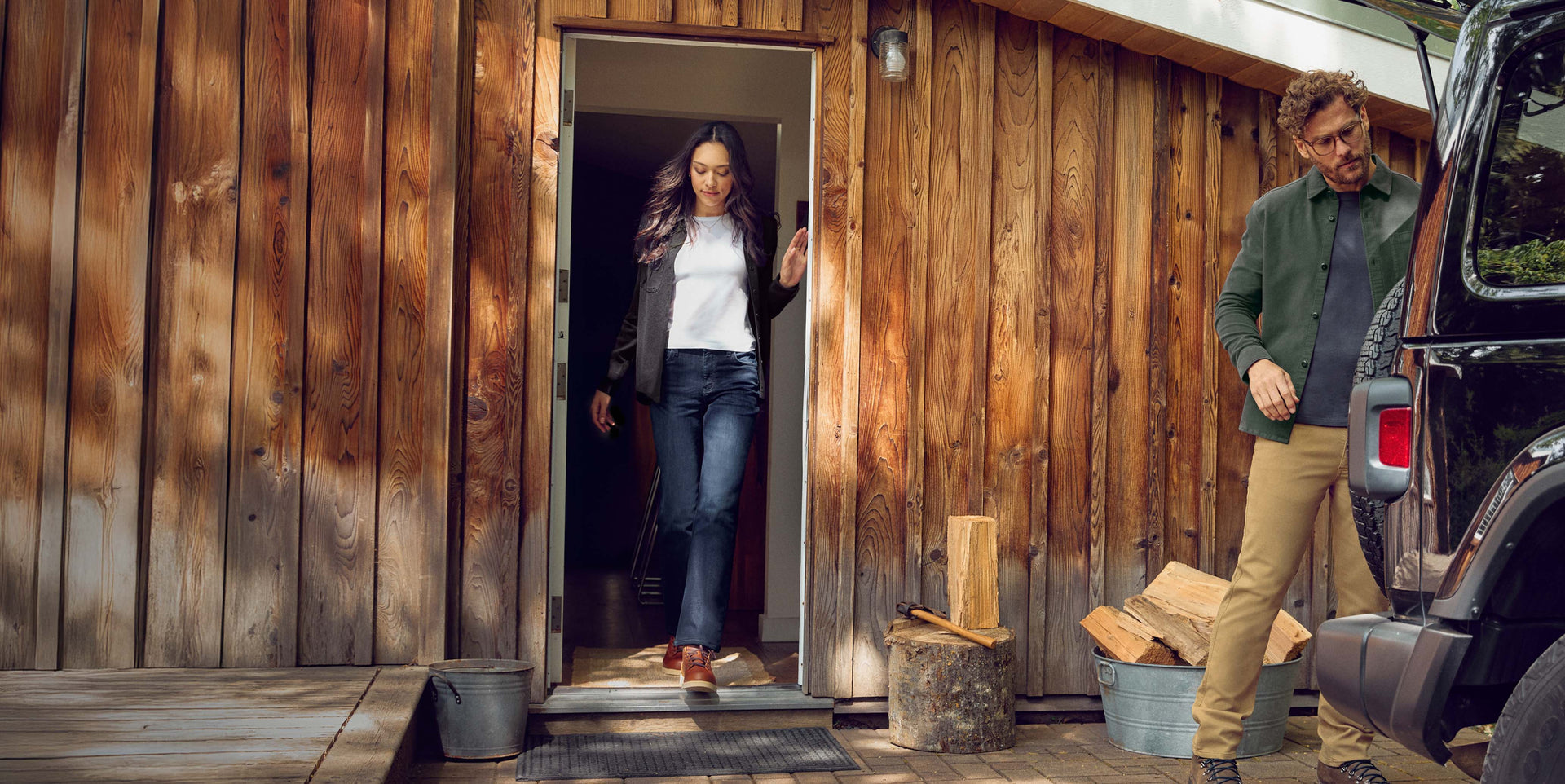 Two people wearing duer denim and duer jeans near a wooden cabin with a vehicle and firewood nearby.