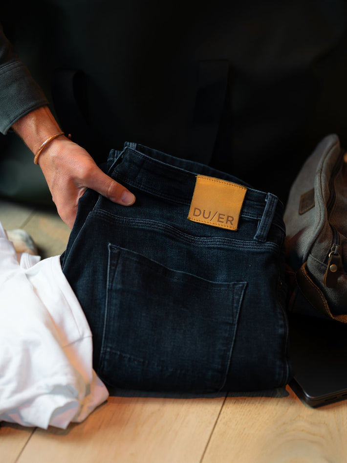 Person holding a pair of dark Blue jeans with a visible DUER Denim brand label on a wooden floor.